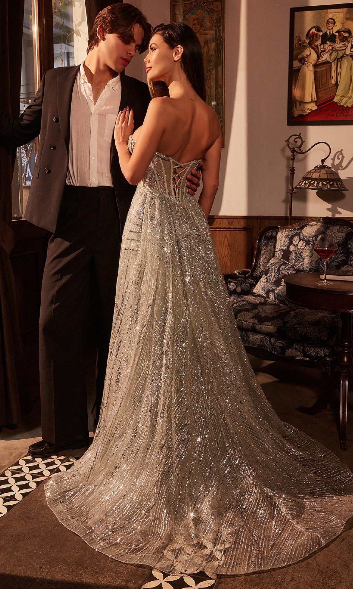 A woman wears the Ladivine CM384 Overskirt Long Glitter Prom Dress, featuring a corset-style bodice, standing beside a man in a black suit and white shirt in an elegant vintage room.